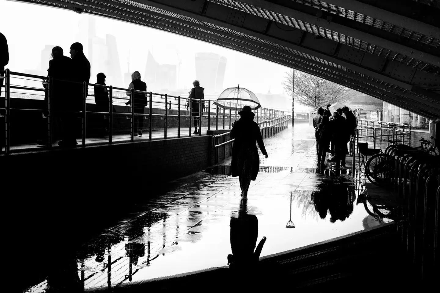 People walking under a bridge in the rain, silhouettes reflected in puddles.