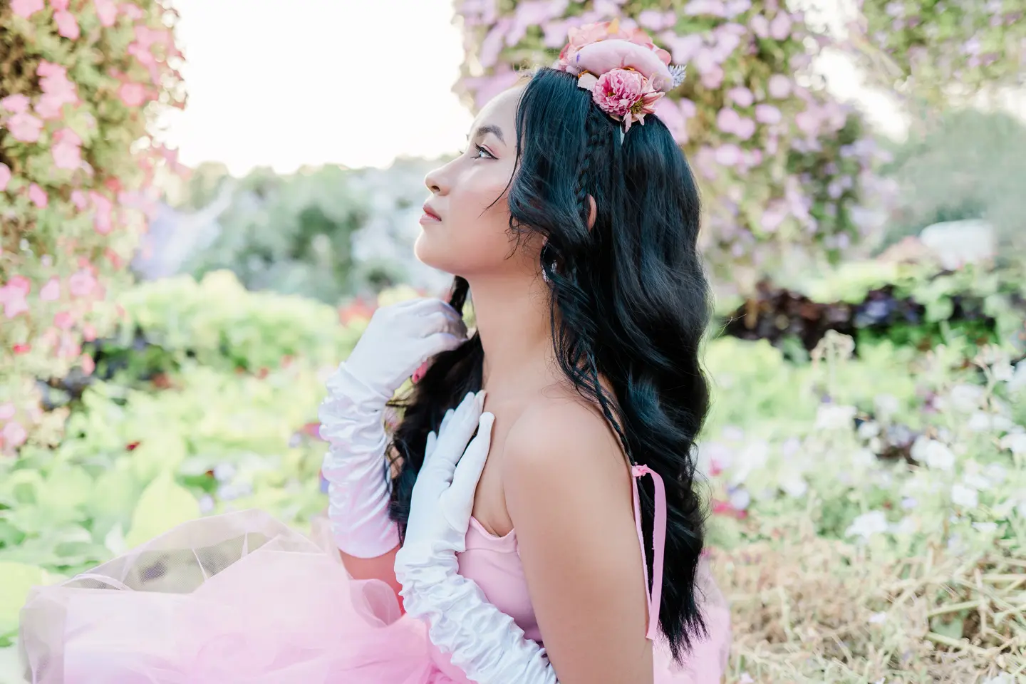 Woman in a pink dress and white gloves is sitting in a rose garden looking to the side.