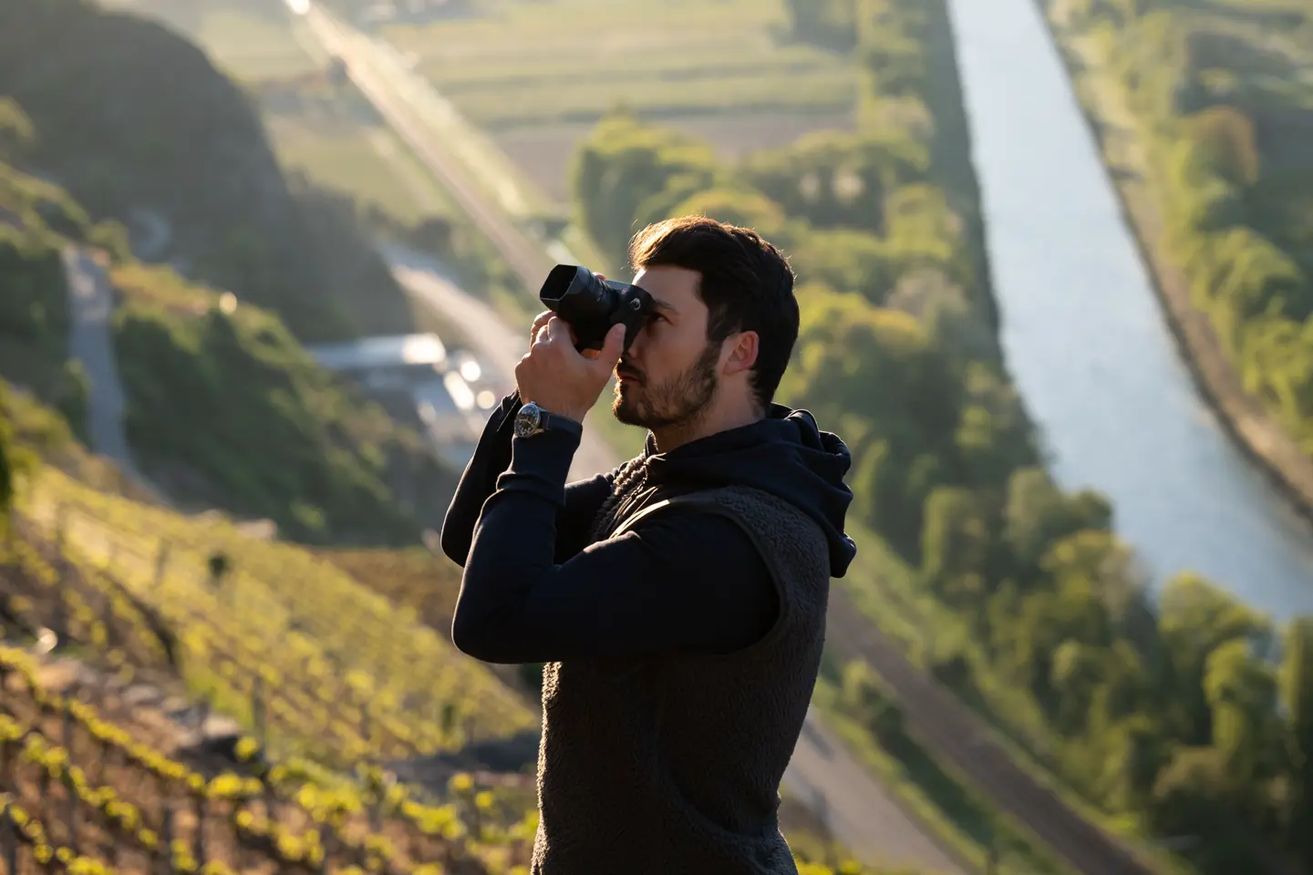 A photographer takes a picture on a sunlit vineyard with a river and trees in the background.