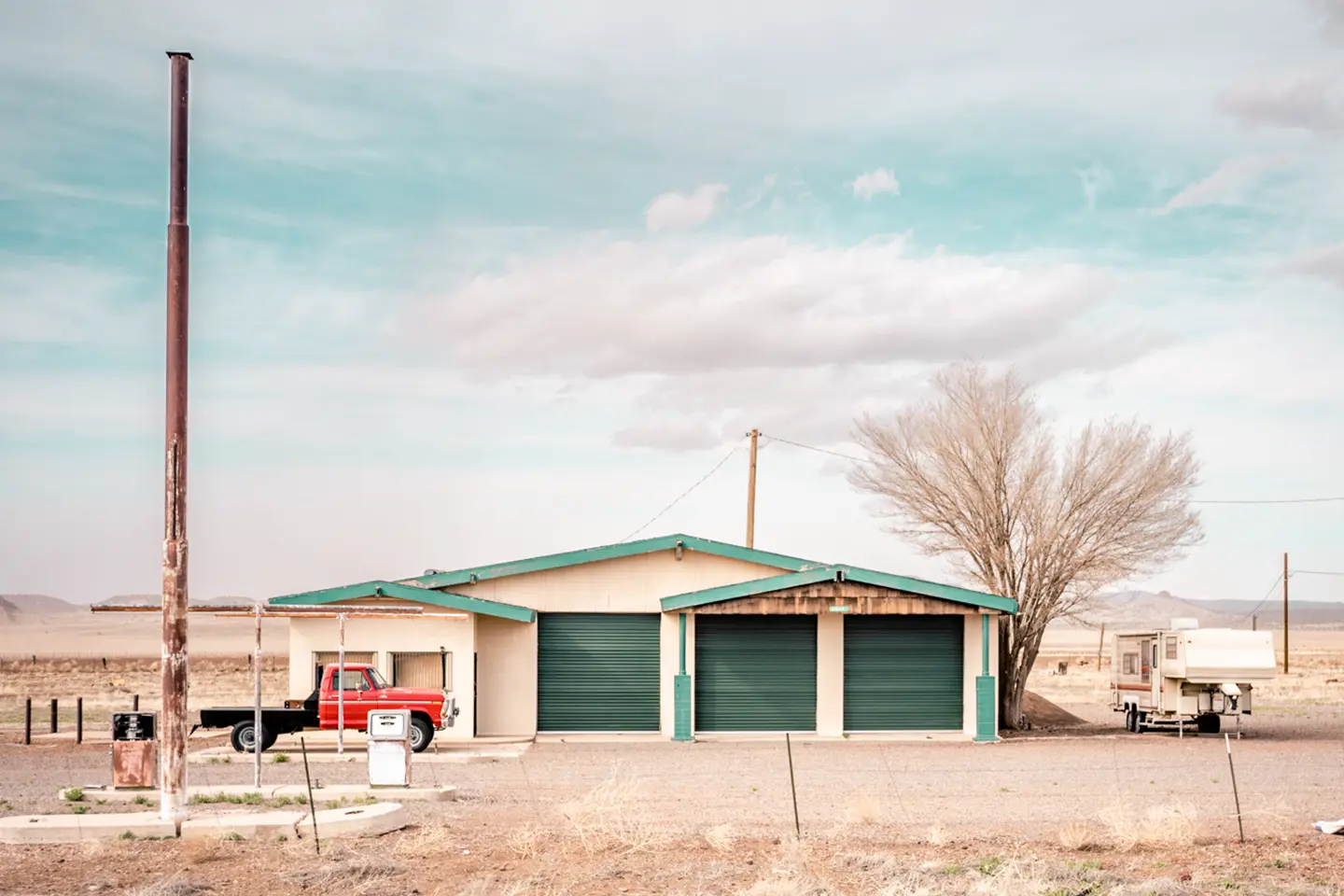 An abandoned gas station in an arid landscape with an old red truck and a caravan.