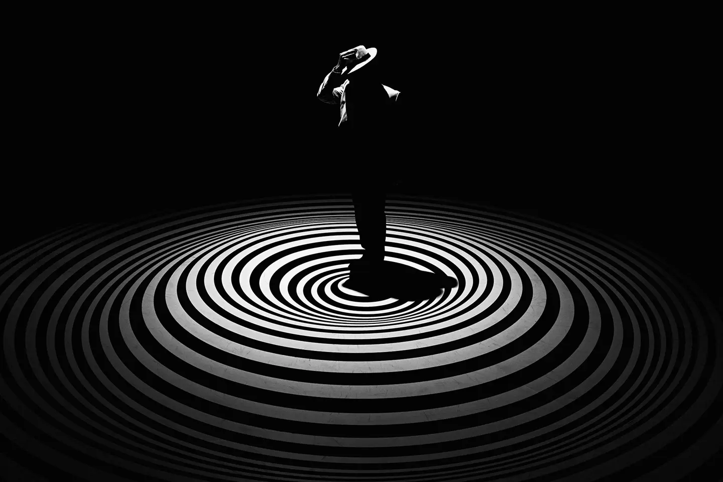 A single person with a hat stands in the centre of concentric circles on a dark background