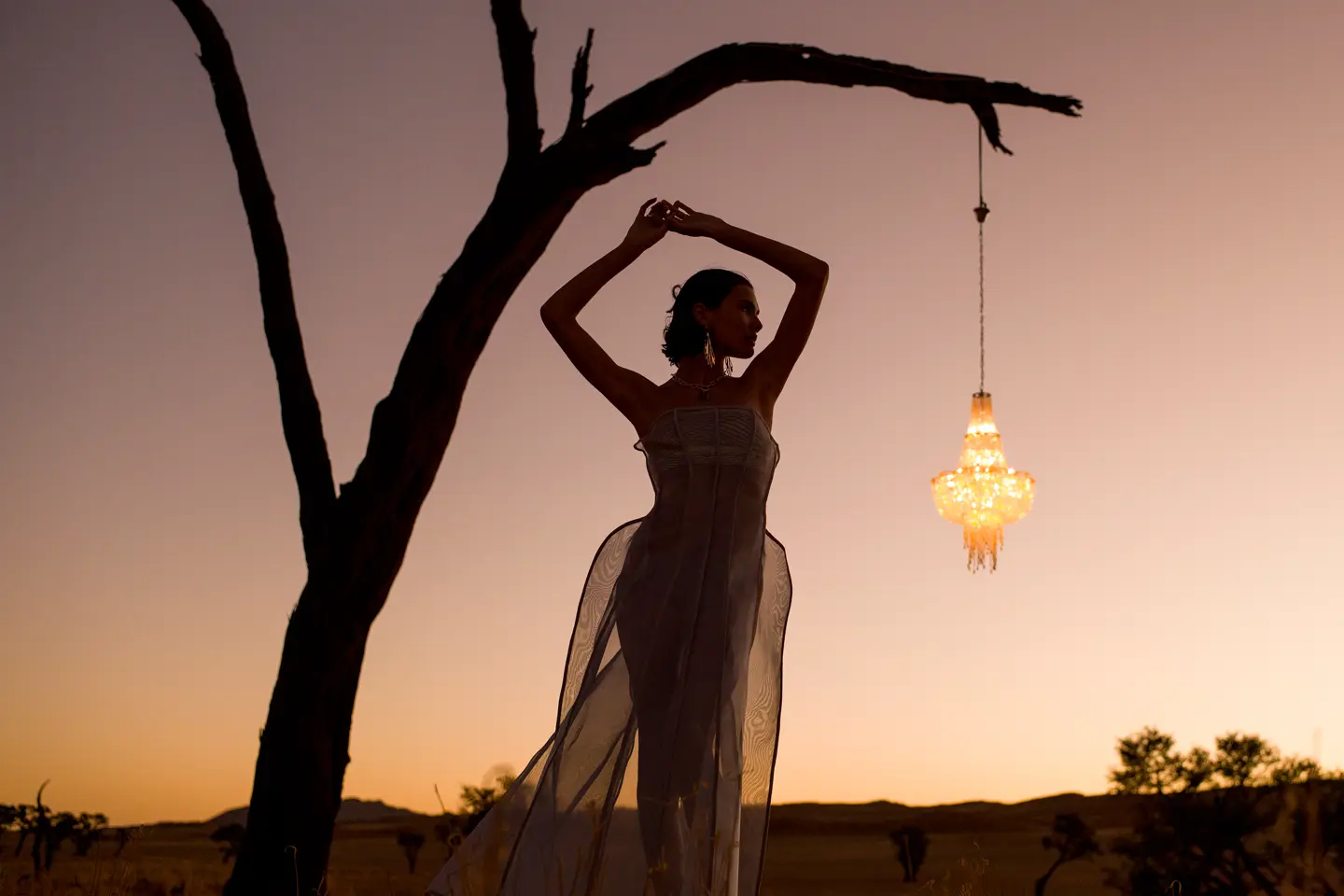 Woman posing under a tree at sunset next to a hanging lantern.