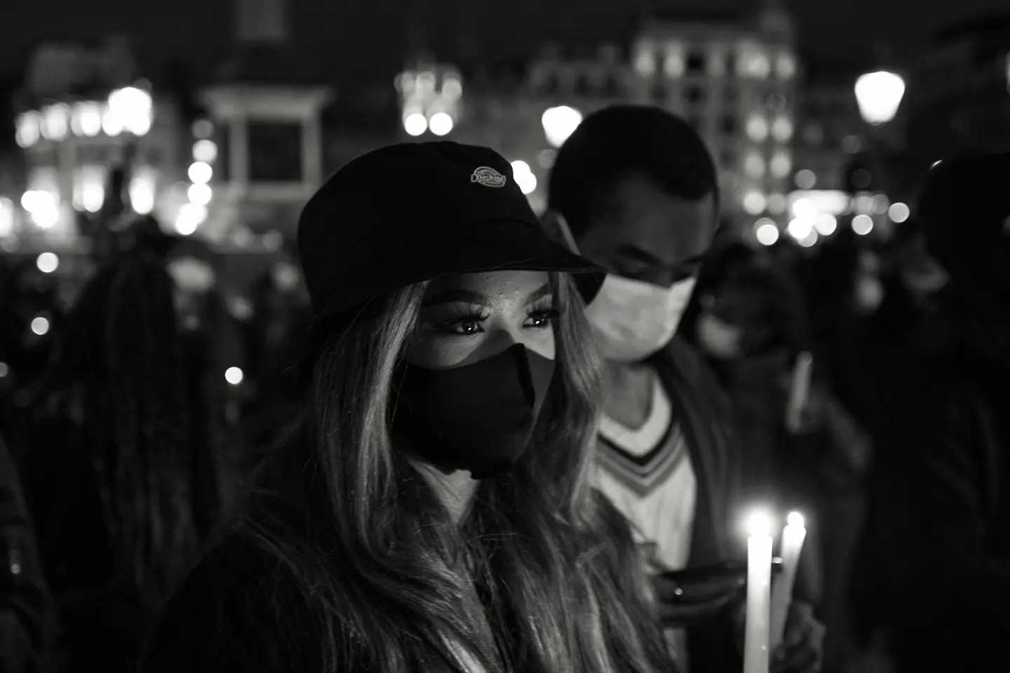 Close-up of a woman and man wearing masks and holding candles in a city at night.