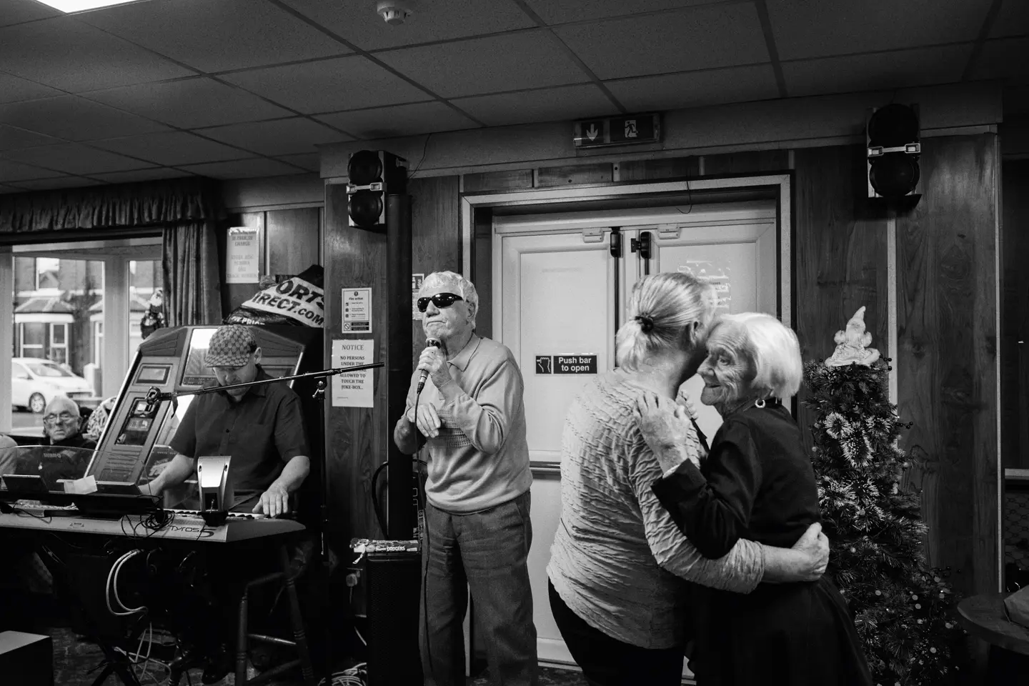 A man sings into a microphone while two older women dance and a man plays the keyboard in the background.