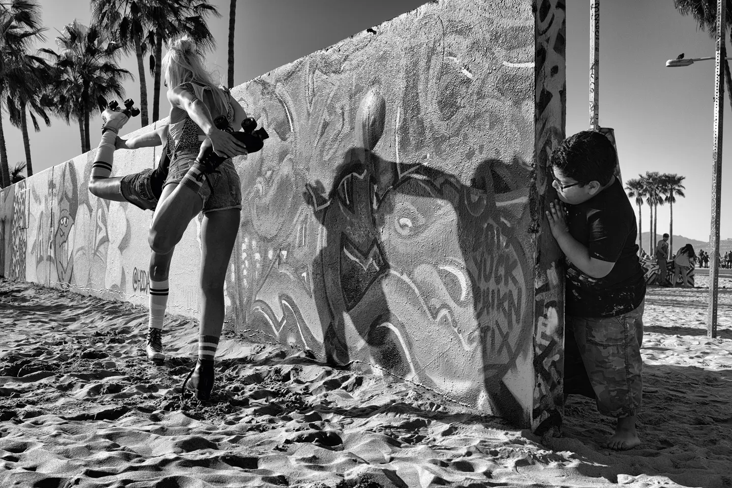 A boy looks behind a graffiti-painted wall at two women on roller skates warming up.