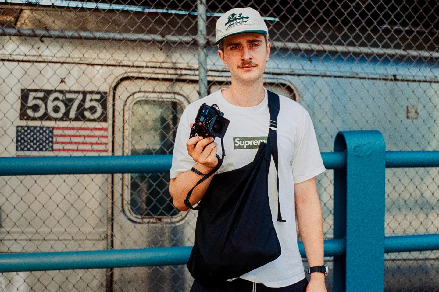 A man with a camera stands in front of a fence and an old train with an American flag painted on it.