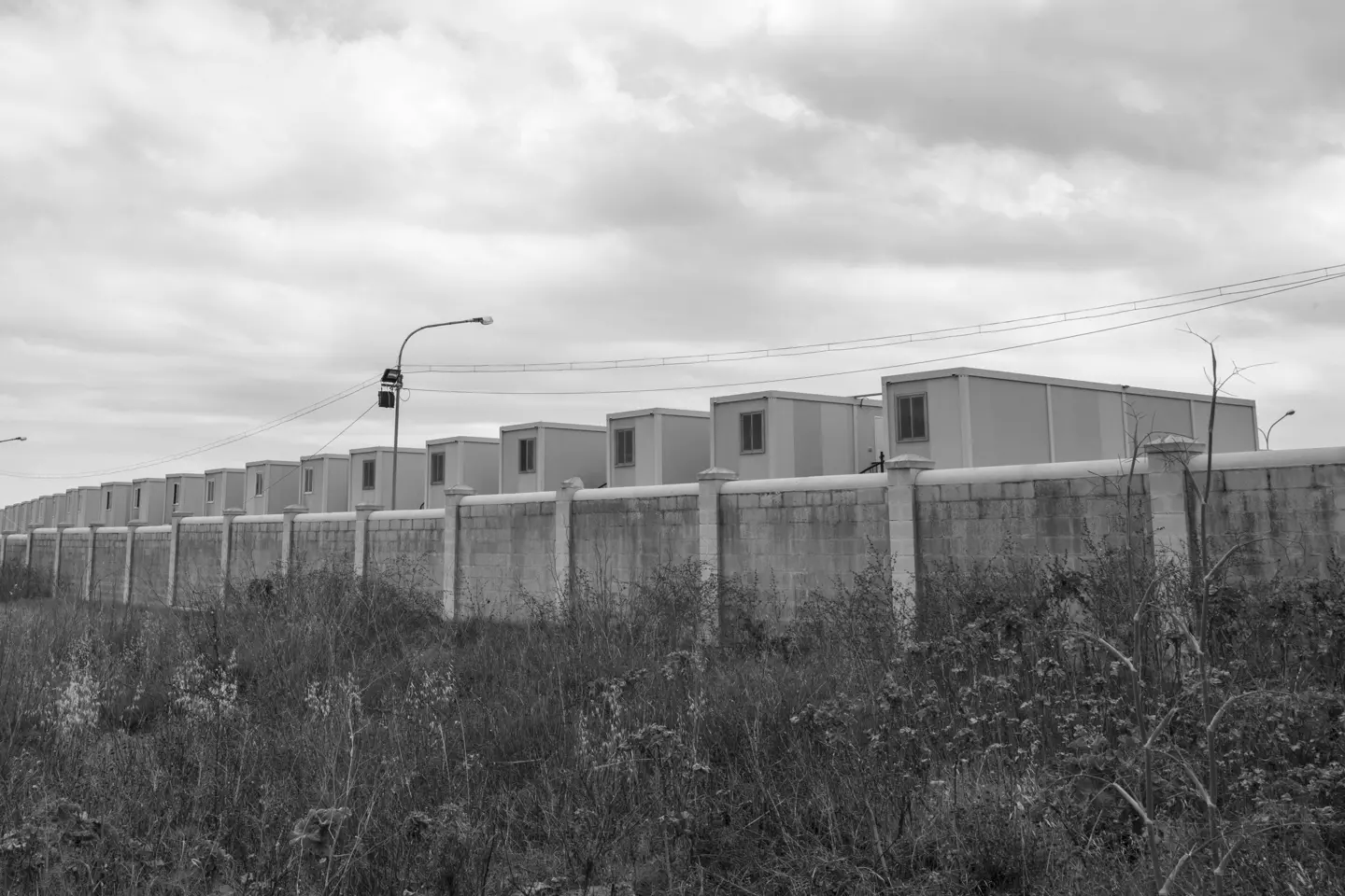 Black and white photo of container houses behind a wall, under a cloudy sky.