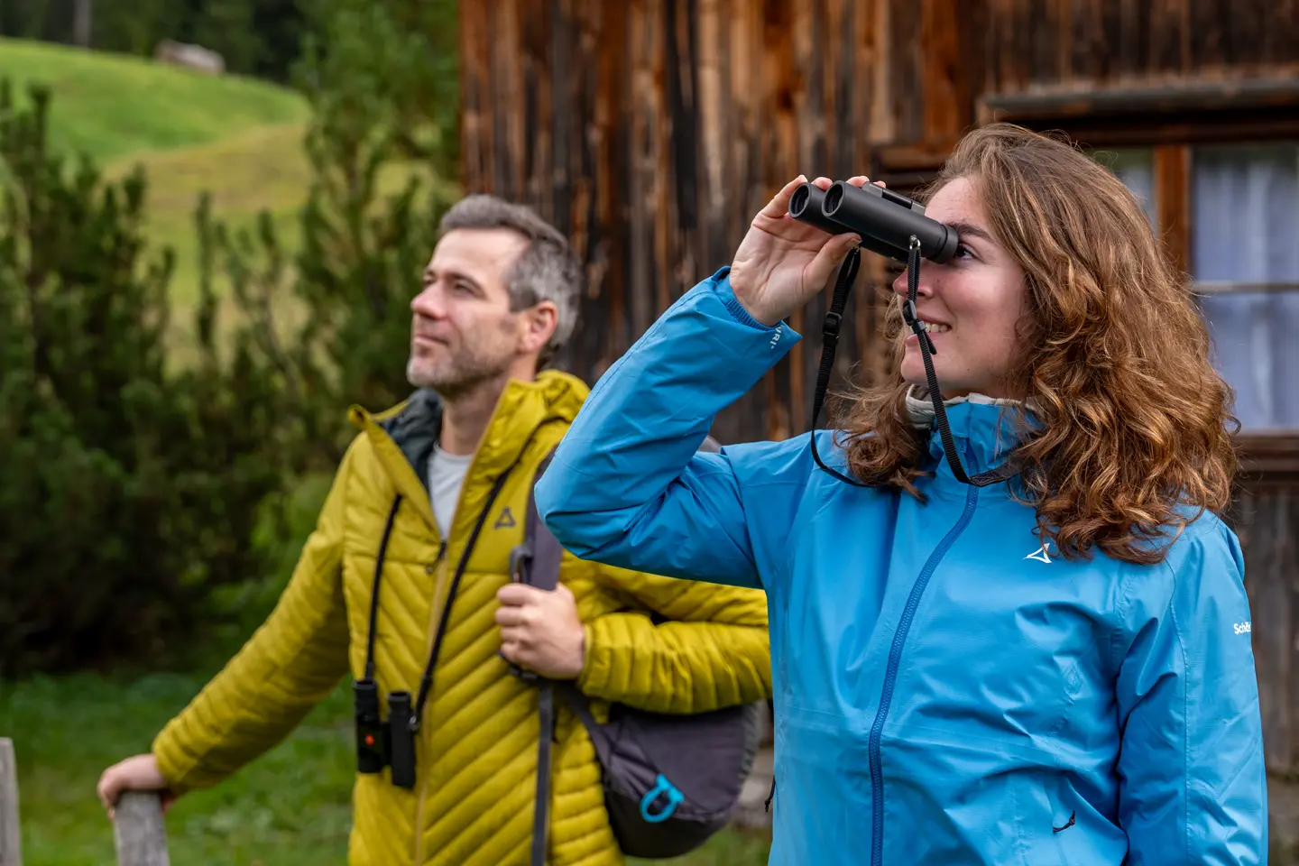 outdoor-hiking-couple-woman-using-leica-noctivid-compact-8x25-binoculars-man-standing-beside-wooden-cabin-green-nature-background