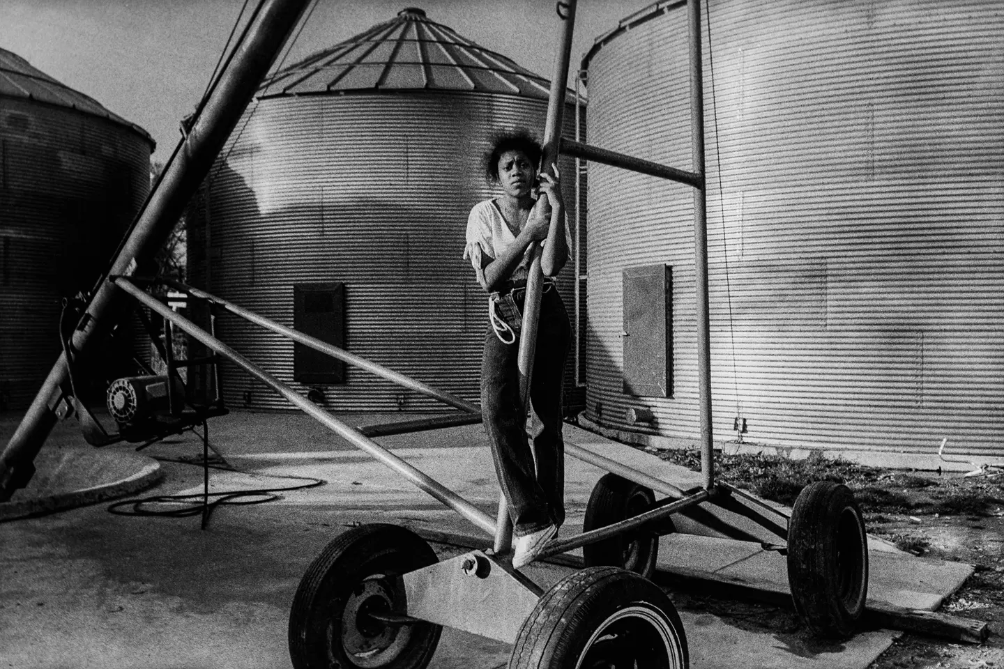 Young girl standing beside farm equipment and grain silos on a family farm in Arkansas, captured in black and white. A documentary photograph reflecting rural life, agricultural heritage, and generational connection.