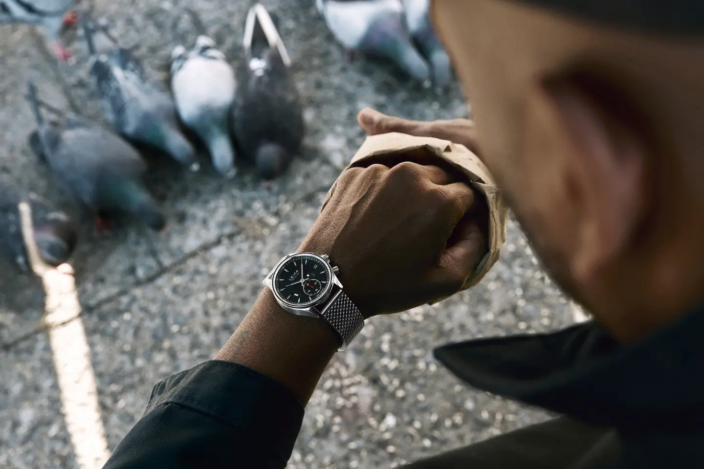 Jason Roman checks his Leica Watch while feeding pigeons in an urban public space, capturing a candid moment of everyday city life and human interaction with wildlife.