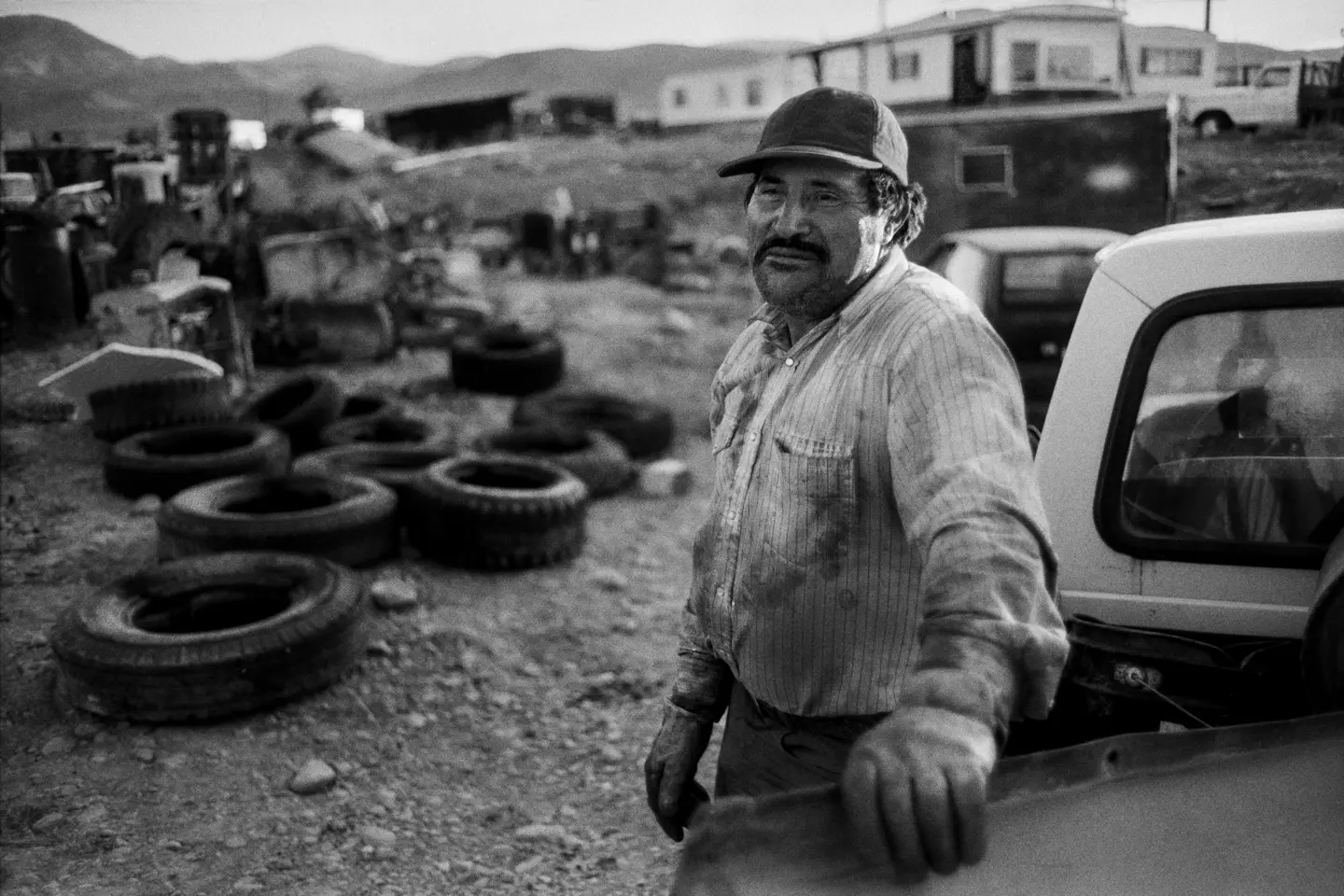 A man in a worn work shirt leans against a pickup truck in a rural scrapyard in Montana. Old tires, machinery, and trailers fill the foreground, with the Rocky Mountains in the distance.