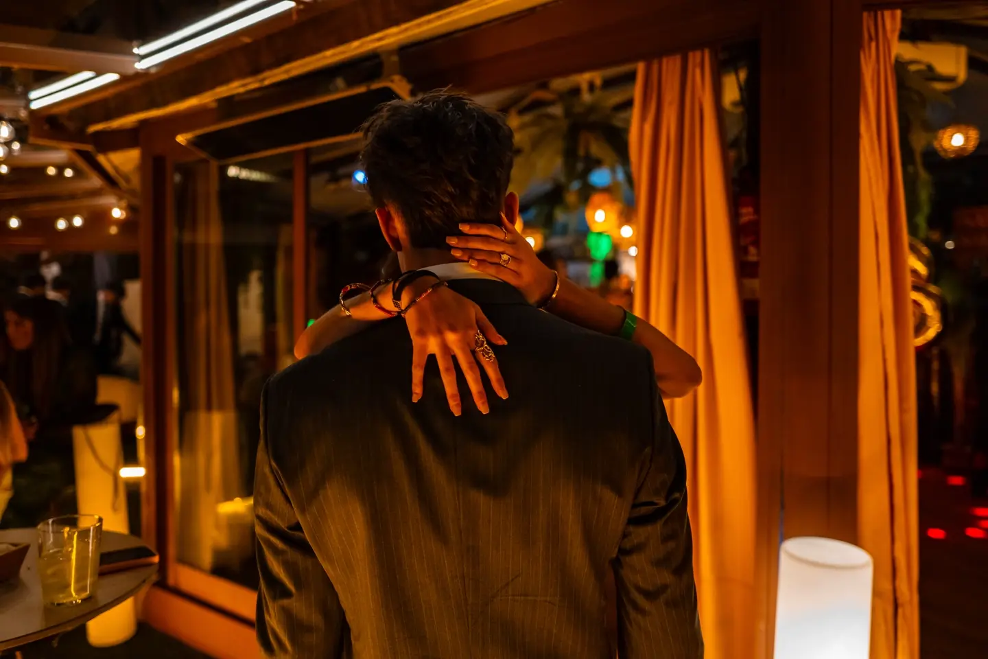 Documentary photograph of a couple embracing at a warmly lit evening celebration, capturing intimacy, human connection, and contemporary social life.