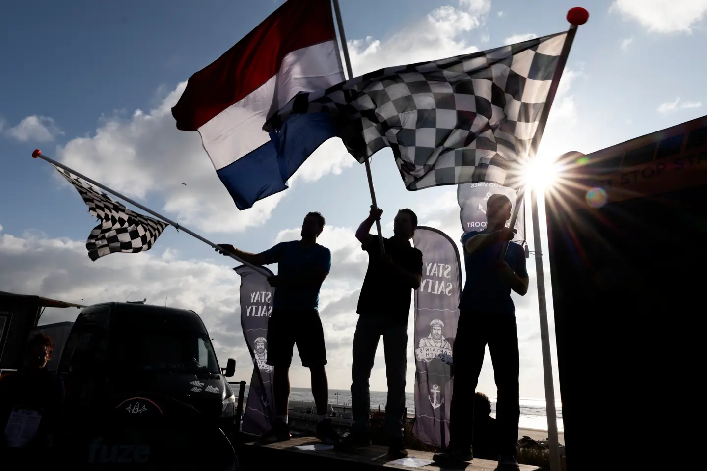 Silhouetted fans wave Dutch and chequered flags at a Formula 1 event in Zandvoort, with bright sunlight breaking through above the seaside circuit.