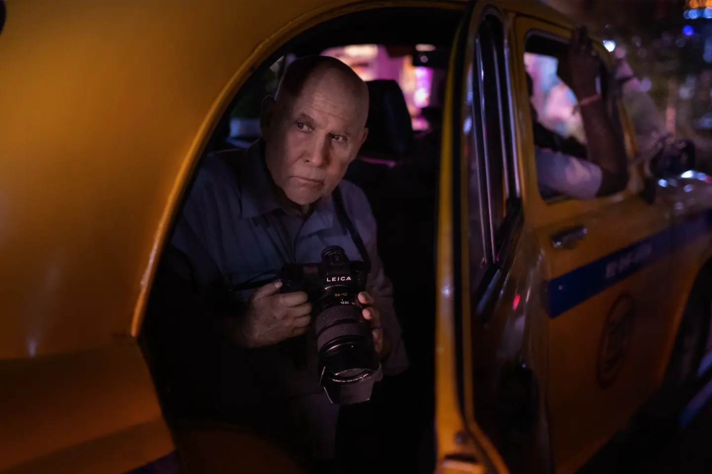 Photographer holding a Leica SL camera while seated inside a yellow taxi at night, illuminated by colorful urban lights.