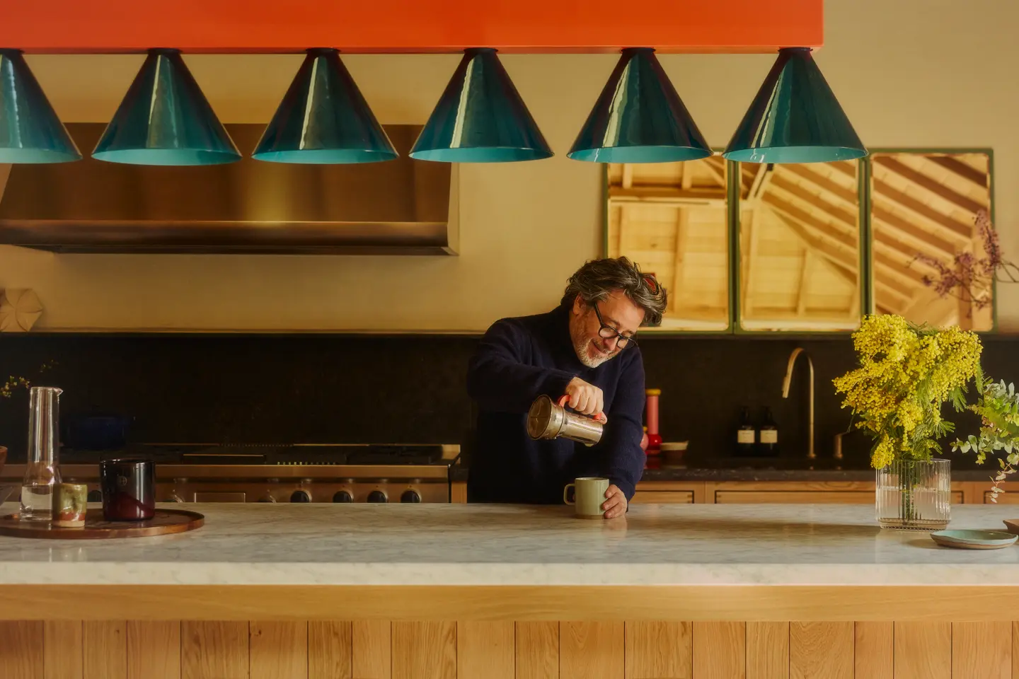 A man pouring a hot drink into a mug behind a large kitchen island in a warmly lit, contemporary kitchen.