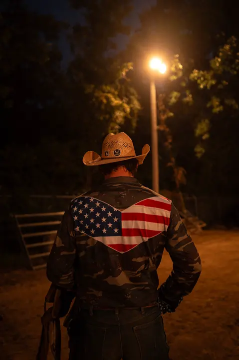 An American Person wearing a cowboy hat and American flag shirt under a streetlight, capturing a quiet nighttime moment in a rural setting.