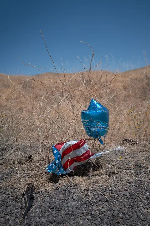 Deflated balloons caught in dry grass beneath a clear blue sky, capturing themes of abandonment, landscape, and everyday visual storytelling.