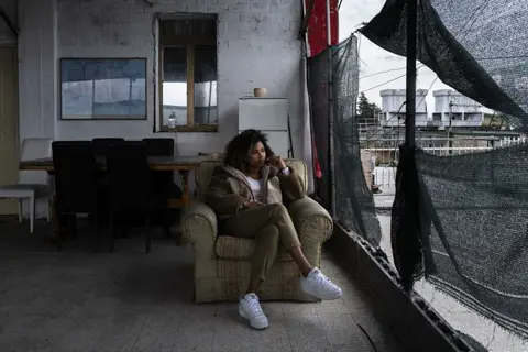 Young woman seated in a modest communal space, capturing contemplation, domestic surroundings, and contemporary urban life.