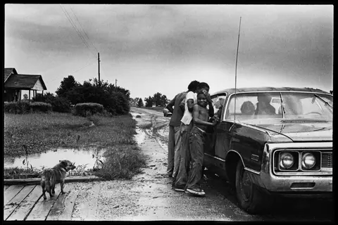 Family members gather beside a car after a summer rainstorm in rural Arkansas. Black-and-white analog photograph from Lisa McCord’s long-term documentary series “Rotan Switch.”