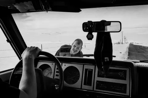 Black-and-white documentary photograph taken from inside a pickup truck, showing a man pushing the vehicle along a snow-covered road in rural Montana, capturing everyday resilience in the American West.