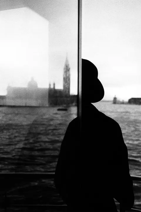 Schwarzweißes analoges Silhouettenbild einer Person am Fenster mit Blick auf Wasser und eine ferne Stadt.