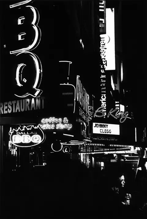 Schwarzweißes analoges Straßenszenenbild mit leuchtenden Neonreklamen und Reflexionen in einer nächtlichen Großstadt.