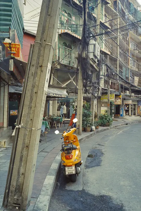 Street scene with a yellow scooter, dense overhead cables and mixed-use buildings, capturing the layered character of everyday urban life.