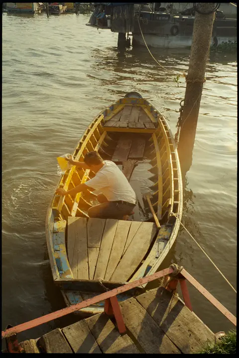 Man bailing water from a weathered wooden boat at a riverside dock, captured in warm evening light and documentary style.