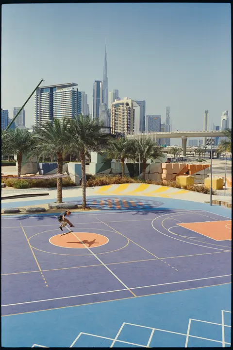 Skateboarder performing a trick on a colorful outdoor court in Dubai, with palm trees and the Burj Khalifa skyline in the background.