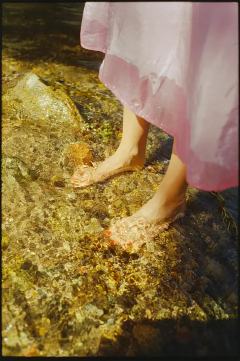 Bare feet standing in clear shallow water beneath a flowing pink dress, capturing sunlight, texture and a quiet summer moment.