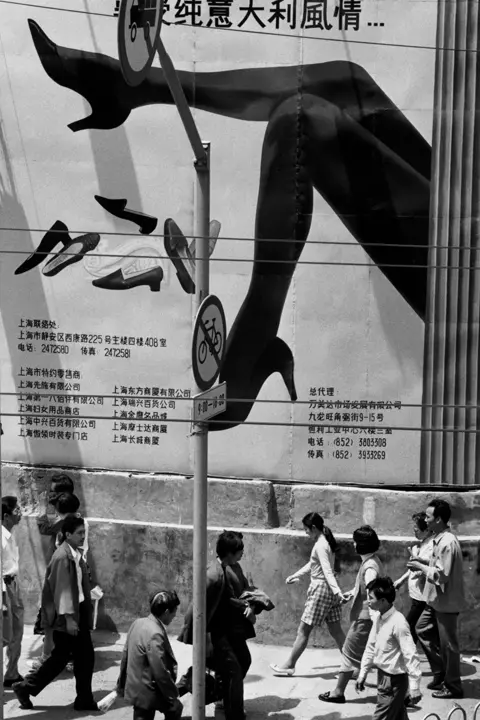 Black-and-white street photograph by Marc Riboud in Shanghai showing pedestrians beneath a large fashion advertisement with oversized legs and shoes.