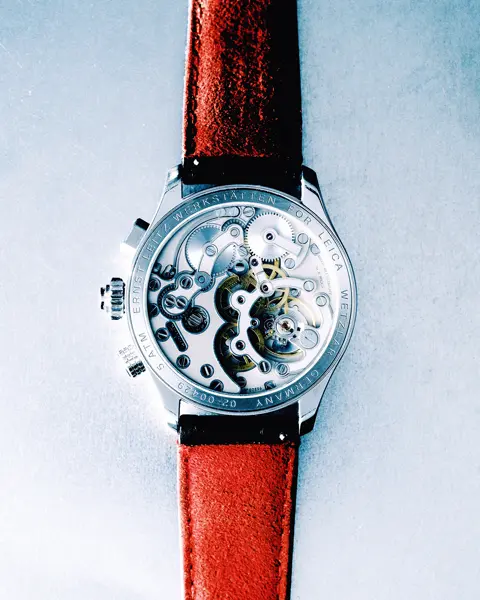 Close-up of a Leica wristwatch with a transparent case back, revealing the mechanical movement and a red leather strap.