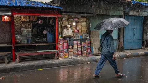 Man walking with an umbrella through heavy rain past a small street bookshop in India, where a bookseller sits among stacks of books.