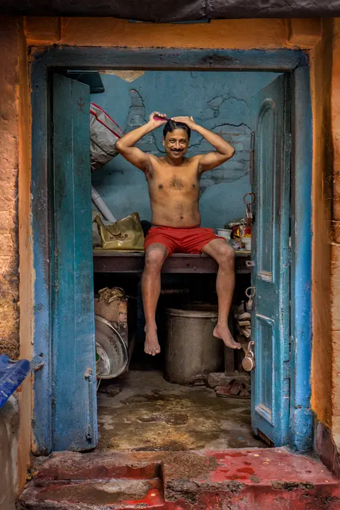 Smiling man in red shorts grooming his hair while seated in the doorway of a modest blue-painted room in India.