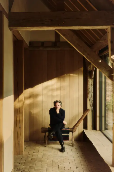 Jay Osgerby dressed in dark clothing sits on a wooden bench in a sunlit hallway with exposed timber beams and wood-paneled walls.