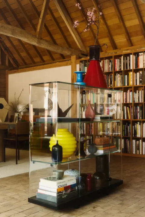 A glass display cabinet filled with colorful vases, decorative objects and books, set against a large bookshelf beneath exposed wooden beams.
