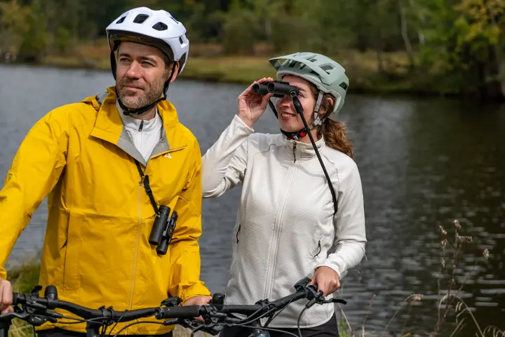 Two cyclists are standing next to a lake and looking through their Leica Noctivid Compact