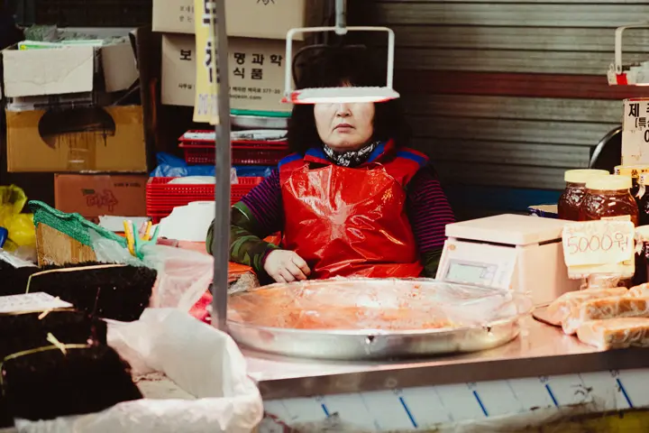 Une commerçante derrière son étal sur un marché, les yeux couverts par un objet suspendu.