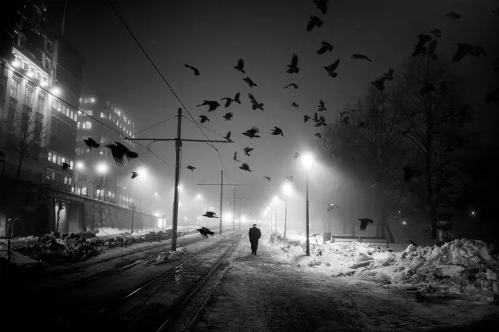 A black and white street scene photograph with a person standing beneath street lights.