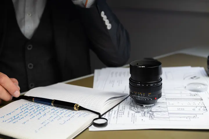 A man is sitting at a desk. There is a Leica lens on the desk.