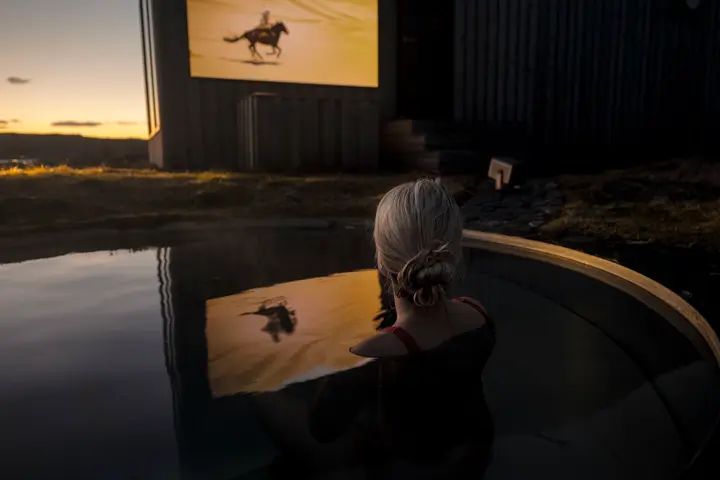 A woman is sitting by the pool in the evening, watching the projection from the Leica Cine Compact 1