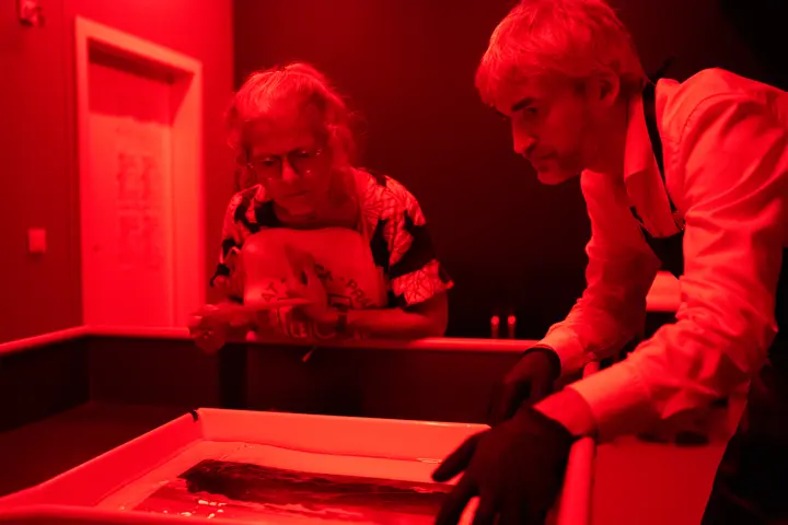 Two people developing a photographic print under red darkroom light.