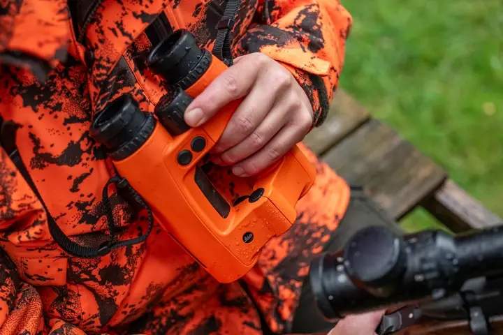Close-up–of–a–hand–holding–orange–Leica–binoculars–beside–a–hunting–rifle