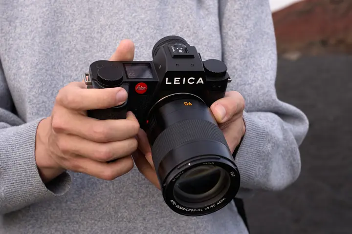 Close-up of a person holding a black Leica SL-System camera with an APO-Summicron-SL 90 mm lens, showing the camera’s top controls, electronic display and front lens design.