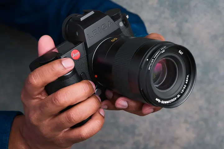 Close-up of a person holding a black Leica SL-System camera fitted with an APO-Summicron-SL 50 mm lens, highlighting the Leica branding, camera controls and large front lens element.
