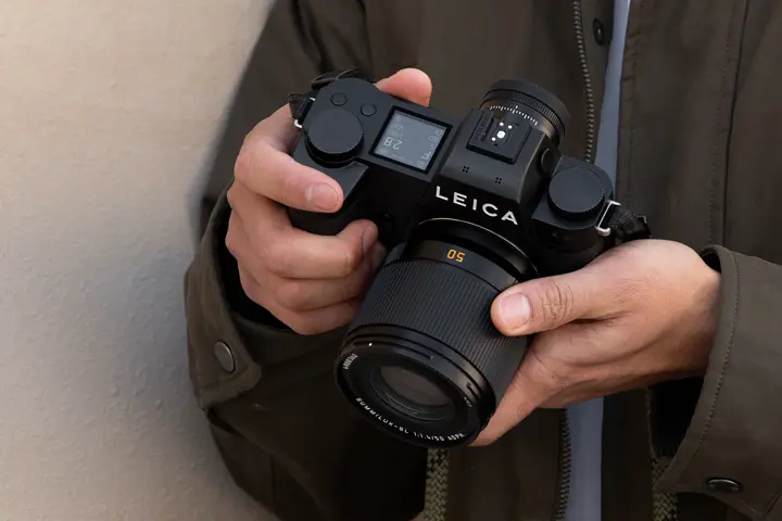 Close-up of a person holding a black Leica SL-System camera fitted with a 50 mm lens, highlighting the camera’s top controls, electronic display and ergonomic grip.