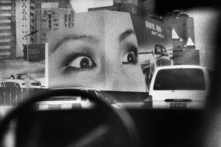 Black-and-white photograph by Marc Riboud showing a busy urban street in China, where cars pass a large billboard featuring a close-up of a woman’s eyes.
