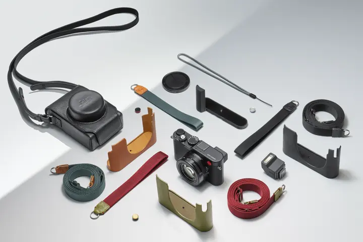 Camera and accessories — Leica compact camera surrounded by straps, cases, grips, a flash and other accessories arranged on a light surface.