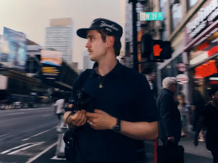 Paul Hepper wearing a Leica wristwatch holds a camera on West 39th Street in New York City, surrounded by motion-blurred traffic and pedestrians.