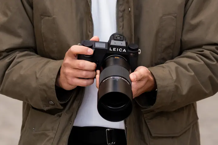 Person holding a Leica SL camera with a telephoto lens, shown in a close-up lifestyle setting.